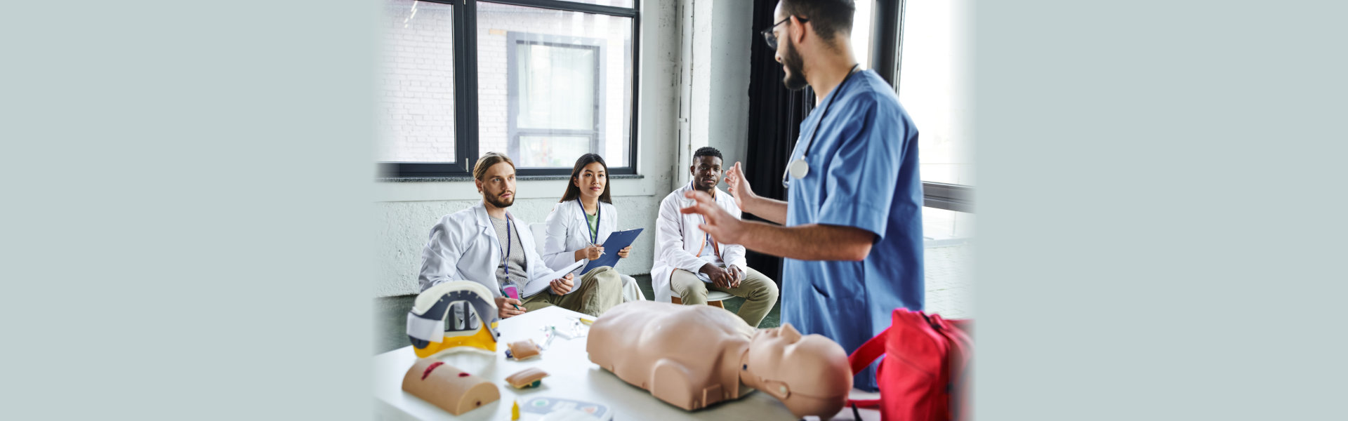 healthcare worker in blue uniform talking to multiethnic students in white coats near CPR manikin
